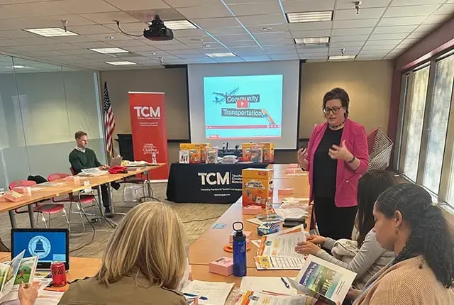 A teach presenting material in front of an audience with a projector screen behind her